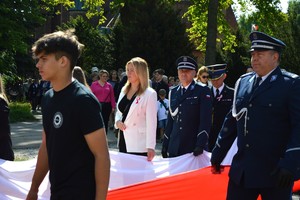 komendant idący wspólnie z gośćmi trzymający długa flagę Polski, biegnącą wzdłuż wszystkich uczestników