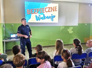 policjant na spotkaniu z dziećmi w tle na wyświetlanym slajdzie napis Bezpieczne Wakacje